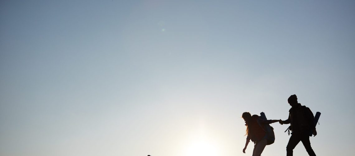 Backlit dark silhouettes of couple hiking in mountains, young man following girlfriends holding hands, side view graphic image