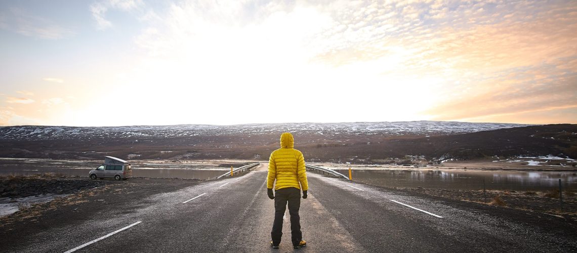 A male wearing a yellow jacket while standing in the middle of an empty road looking in the distance